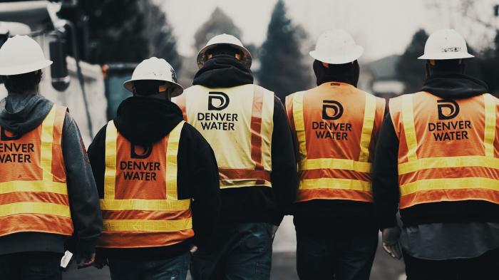 Five men seen walking away from the camera, lined up shoulder to shoulder, with Denver WAter safety vests and hard hats. A team, together, the job complete.