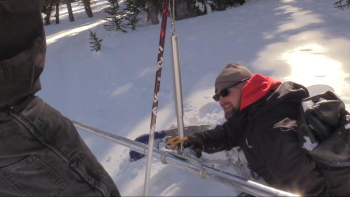 A man lays in the snow taking a measurement by peering at a device holding a metal pole suspended in mid-air. 