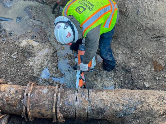 A Denver Water worker wearing a safety vest, helmet and gloves holds a saw that's cutting into an old, exposed pipe laying in the mud and dirt. 