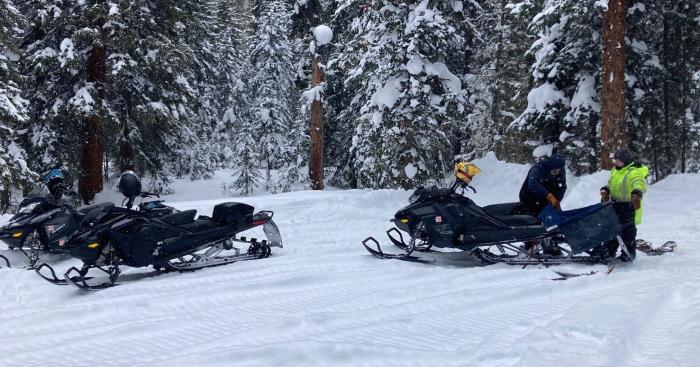 Two men crouch over a snowmobile, stuffing equipment into storage sacks for a trip into the backcountry. There are three snowmobiles in the pictures, and tracks in the snow.