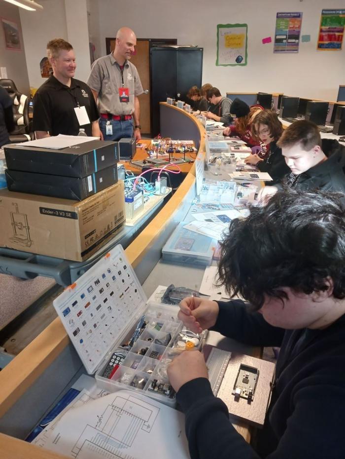 A line of students sitting at desks, heads beant in concentration, as they wire controllers together, while two men look over the classroom smiling proudly.