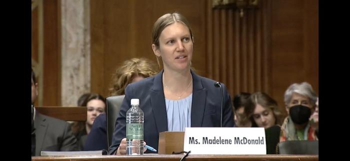 A woman dressed in a suit sits at a long table looking toward the camera. Her name, Ms. Madelene McDonald, is on a name card in front of her, behind her people sit, listening.  