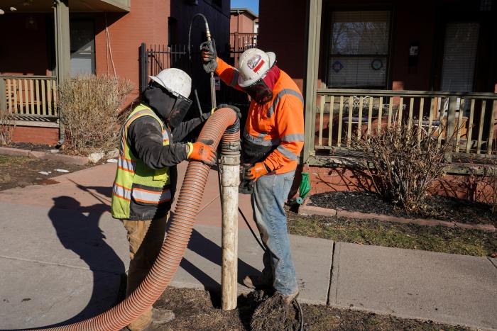 Two workers in construction gear use a power washing hose and a long, orange hose to create a hole in the ground to inspect a water line.
