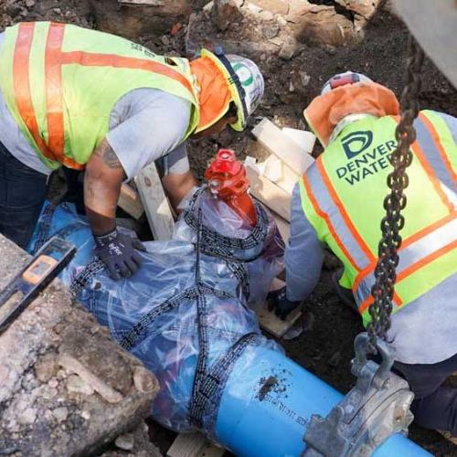 Two men at work, wearing Denver Water yellow safety vests, hard hats with a blue pipe running next to them in the ground.
