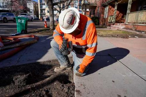 A man wearing a white hard hat and orange safety jacket shines a flashlight down a hole in the dirt to check the service line material.