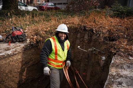 A worker stands in a hole dug in the ground, smiling, holding a coil of copper pipe.