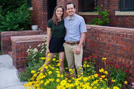 A man and woman stand in front of a home's low brick front all, with yellow flowers at their feet.