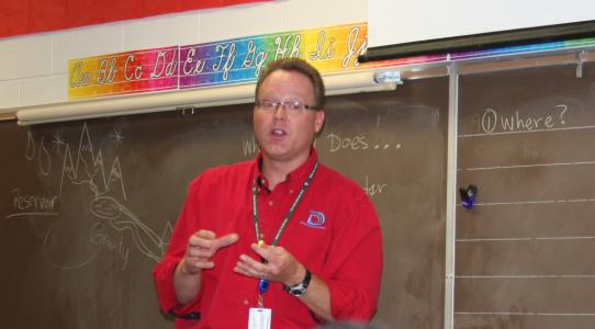 A man in a red shirt with a Denver Water logo stands in front of a chalkboard.