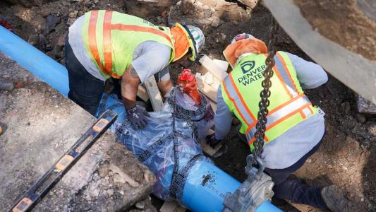 Two men at work, wearing Denver Water yellow safety vests, hard hats with a blue pipe running next to them in the ground.