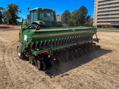 A huge green machine with a cab that looks similar to a combine rolls across brown dirt, with tall buildings in the background.