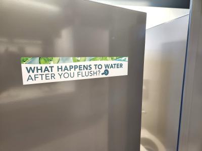 A stainless steel bathroom stall door is slightly open, a reflected image of a toilet seat is seen on the wall. On the door is a sign that asks: "What happens to water after you flush?"