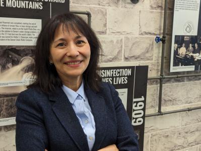 Dark-haired woman in a blue shirt, dark navy suit jacket smiling in front of a stone wall with posters from a timeline.