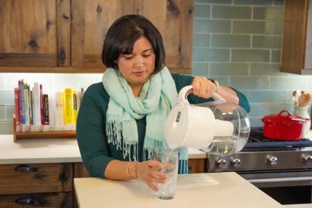 A woman stands at a kitchen counter pouring water from a pitcher and water filter into a glass to drink.