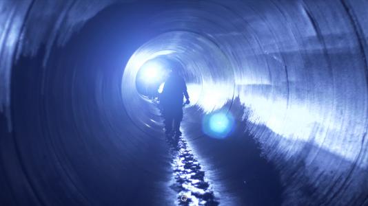 People holding flashlights walk through a huge pipe that looks like a tunnel.