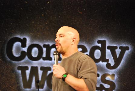 A bald man holds a microphone in front of the Comedy Works sign.