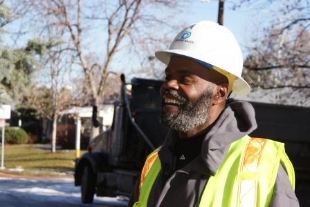 A man in a Denver Water hard hat and yellow safety vest laughs.