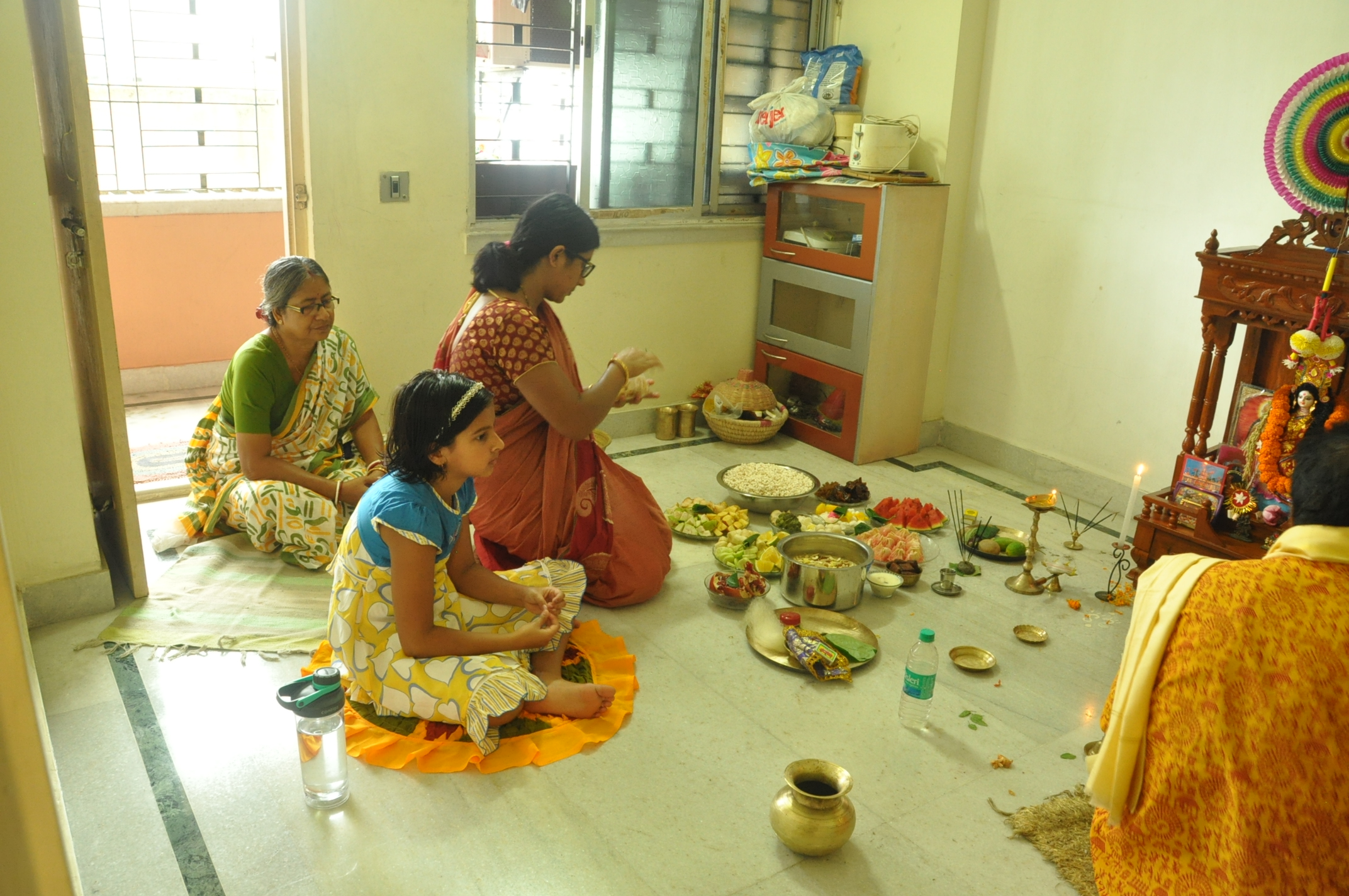 three women sit on a floor filled with bowls and plates of offerings for a ceremony.