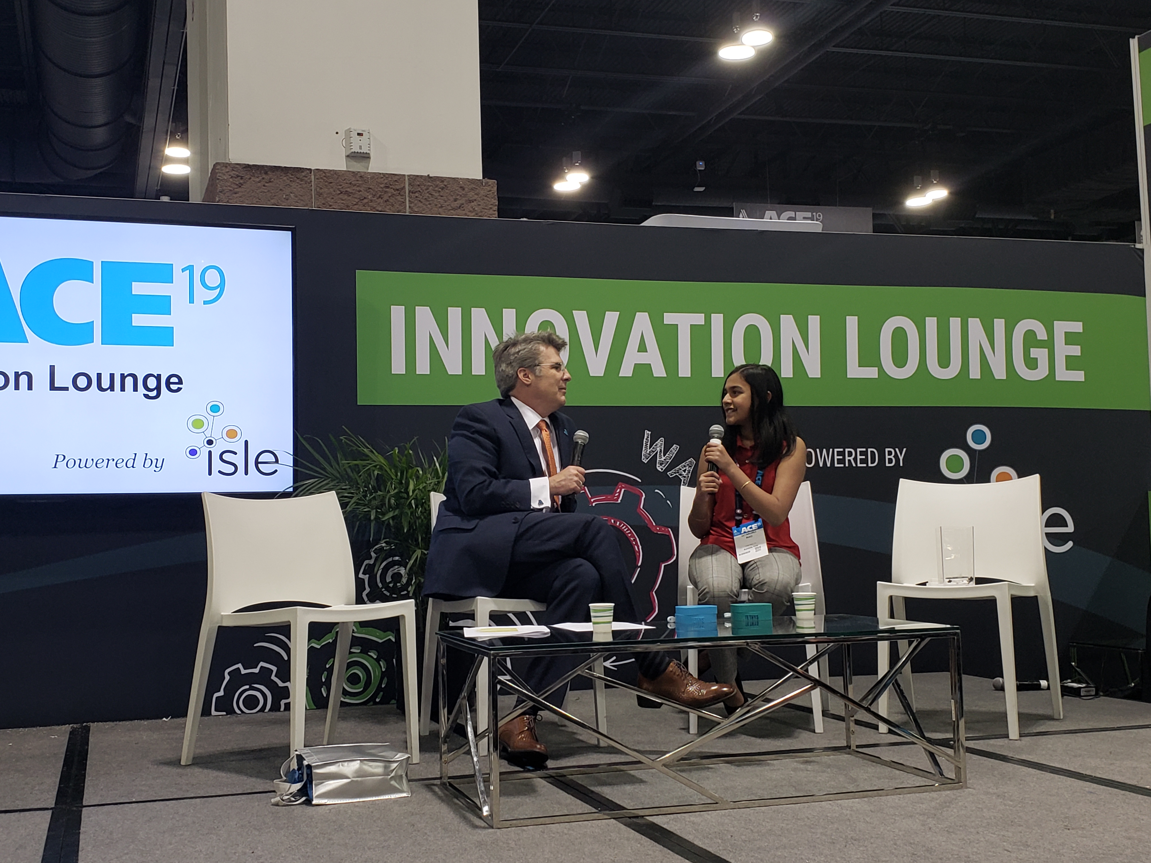 A man and a young woman sit on a stage, with the sign "Innovation Lounge" behind them.