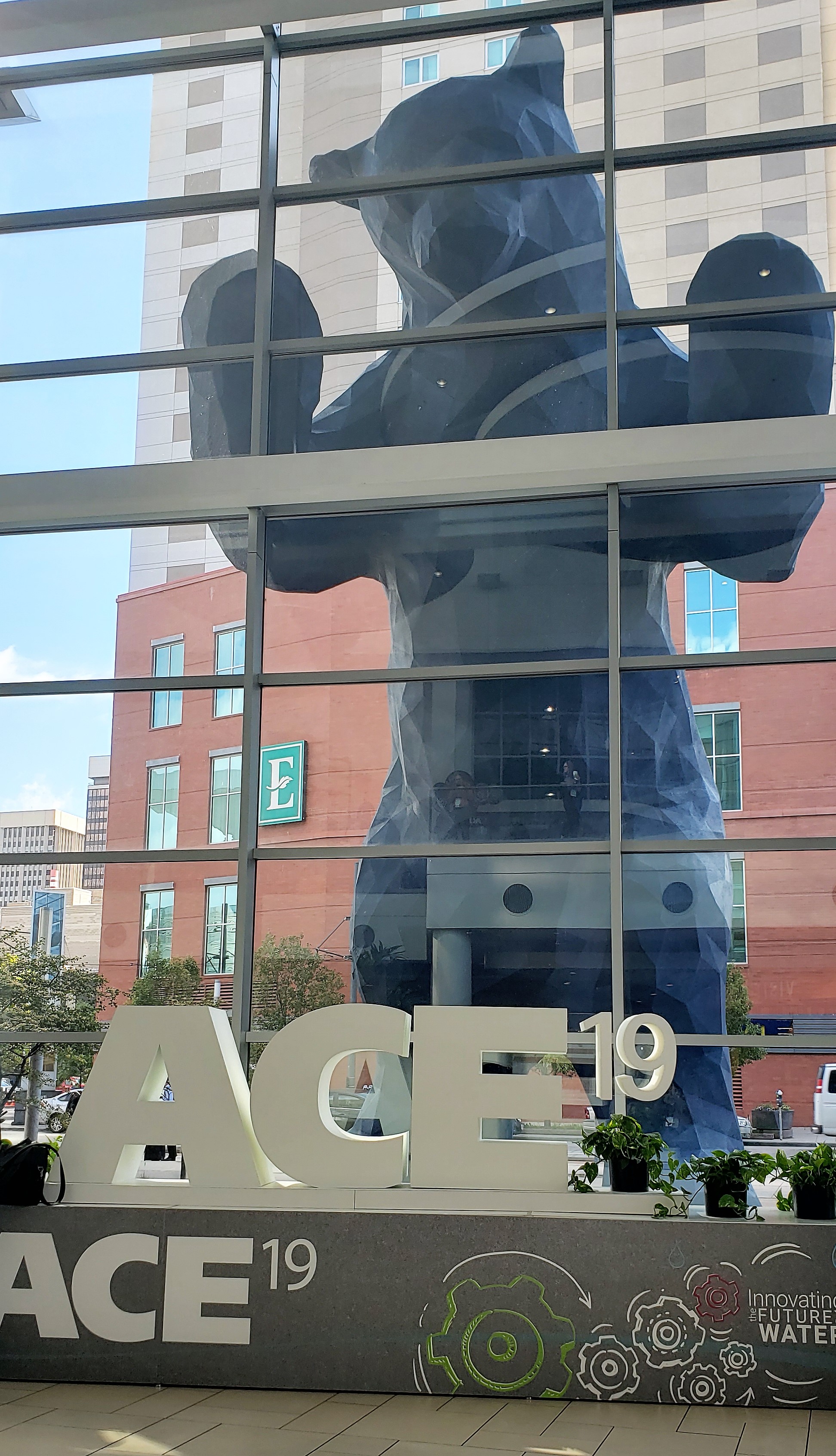 The city of Denver's iconic blue bear statue peers through the glass of the Colorado Convention Center at an information table set up for ACE19.