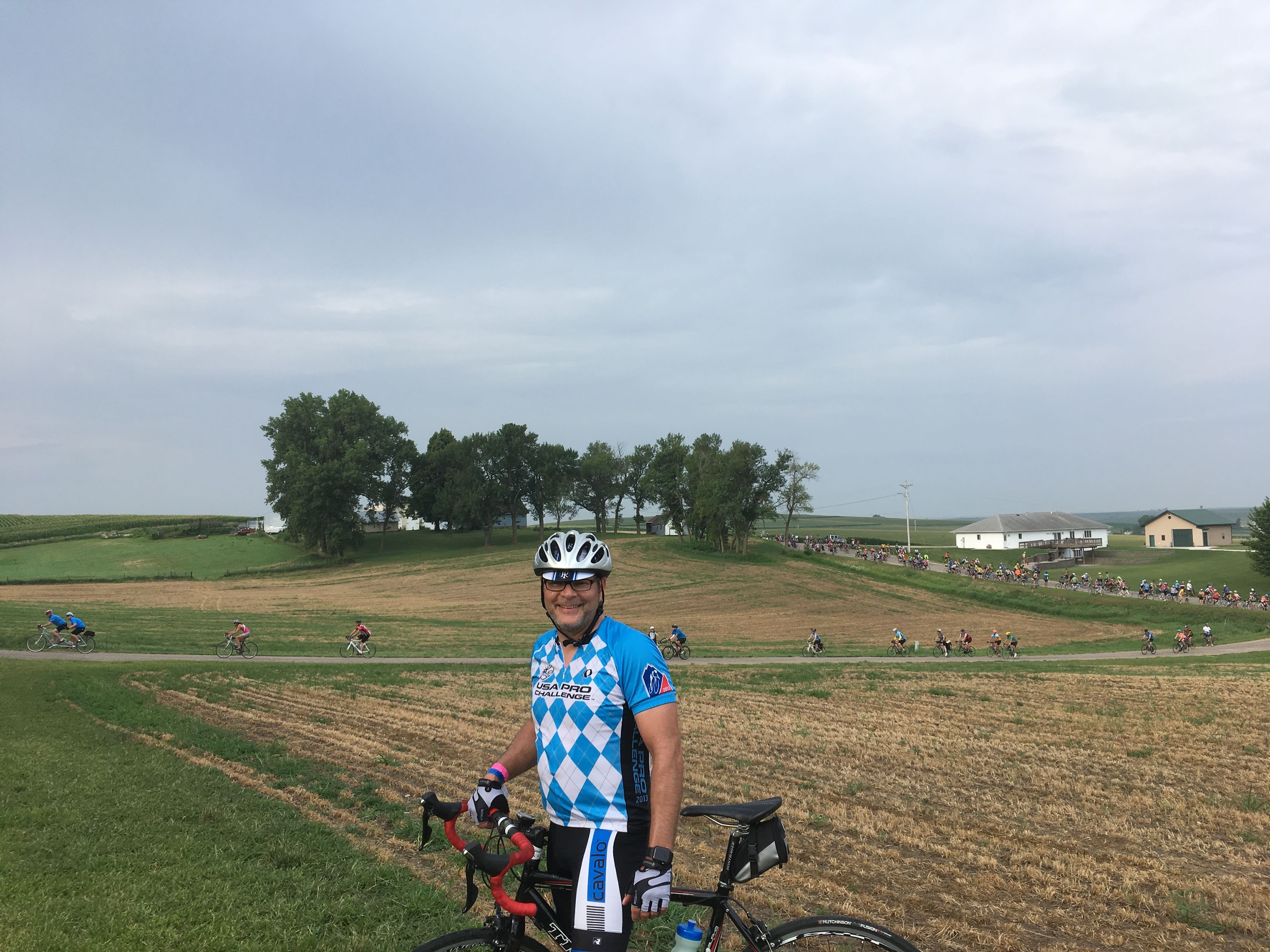 Adam Hutchinson is centered in the picture, showing fields behind him and bicycle riders in the distance.