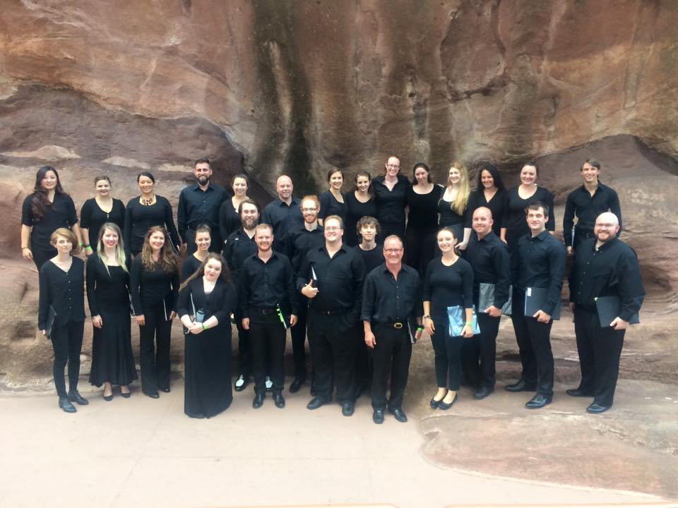 A group of people in black shirts and pants or skirts, smiles before a wall of rock.