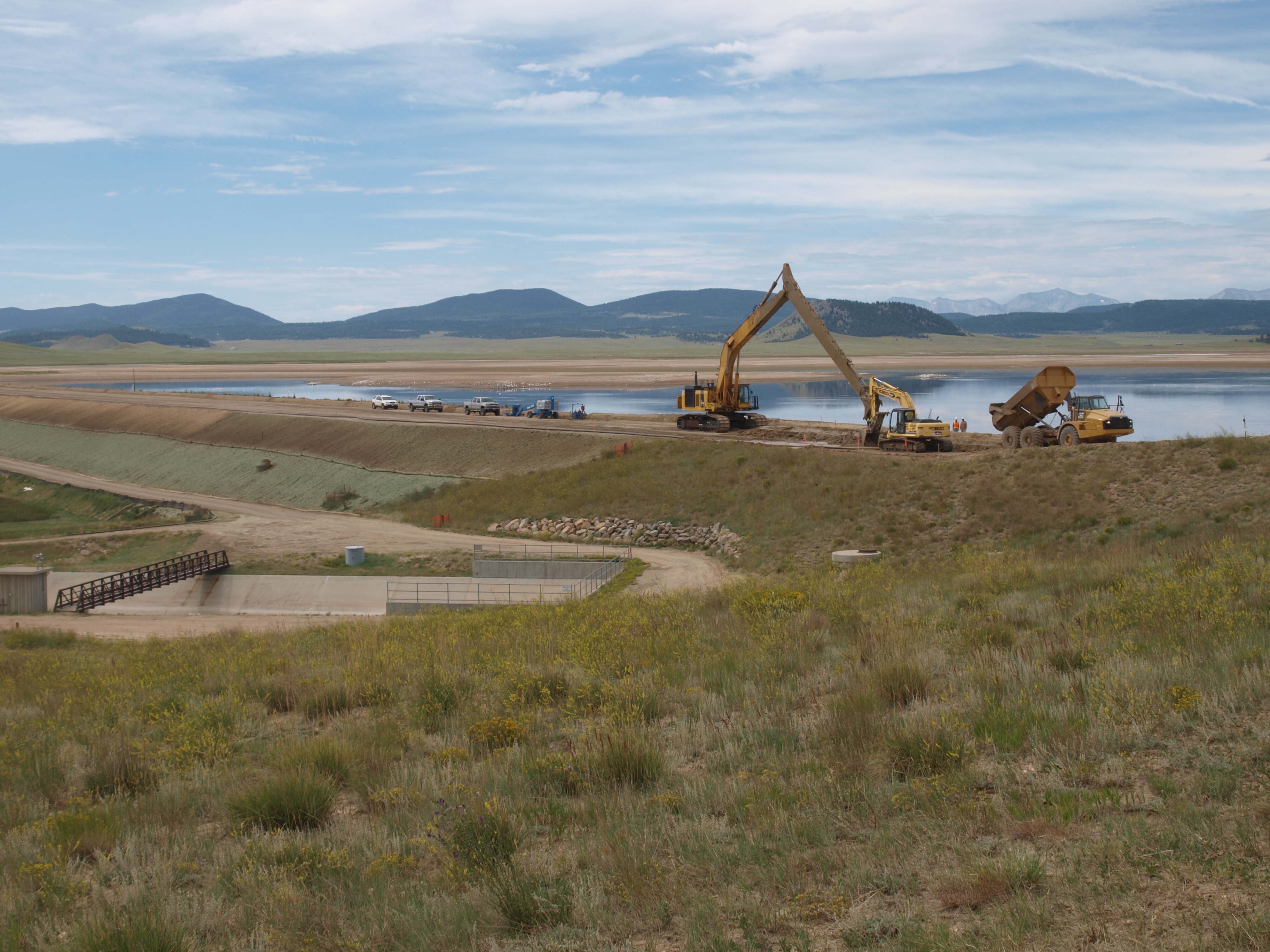 An excavator and heavy truck sits atop a dam with mountains in the distance.