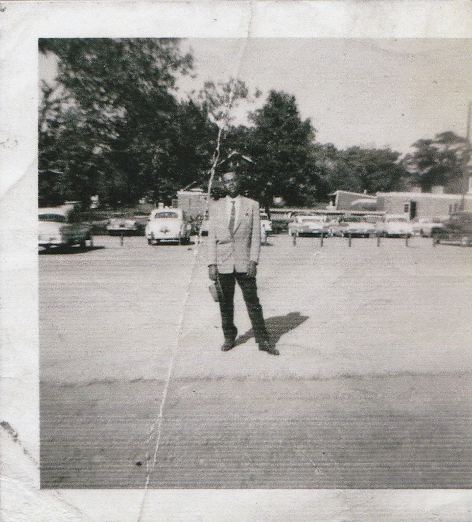 A black man in a suit setands in a parking lot, holding his hat in his right hand.