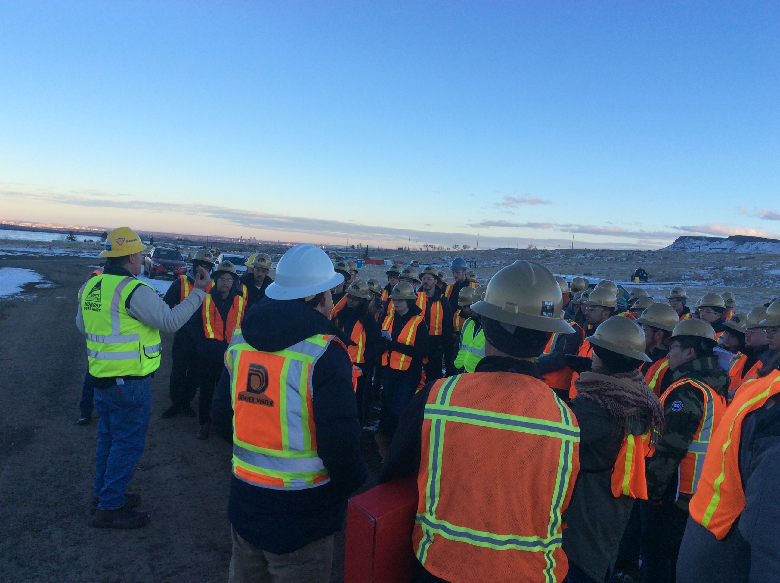 A crowd of students, all wearing hardhats and safety vests, listens to a man in a hard hat talk.