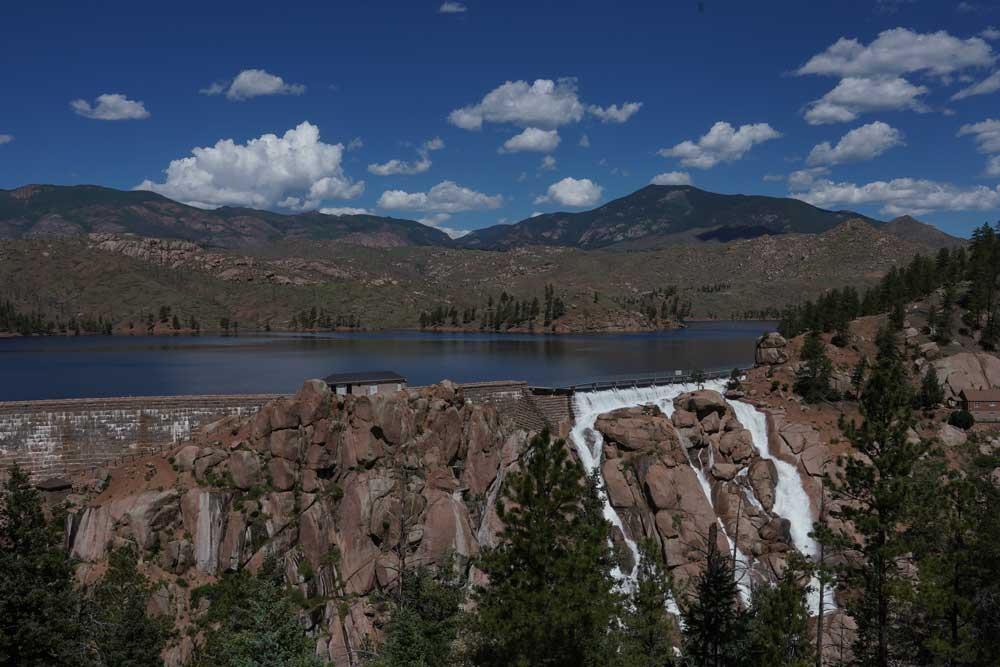 White water spills down a wall of red boulders, withe the flat blue reservoir in the background under mountains and a cloud-dotted blue sky.