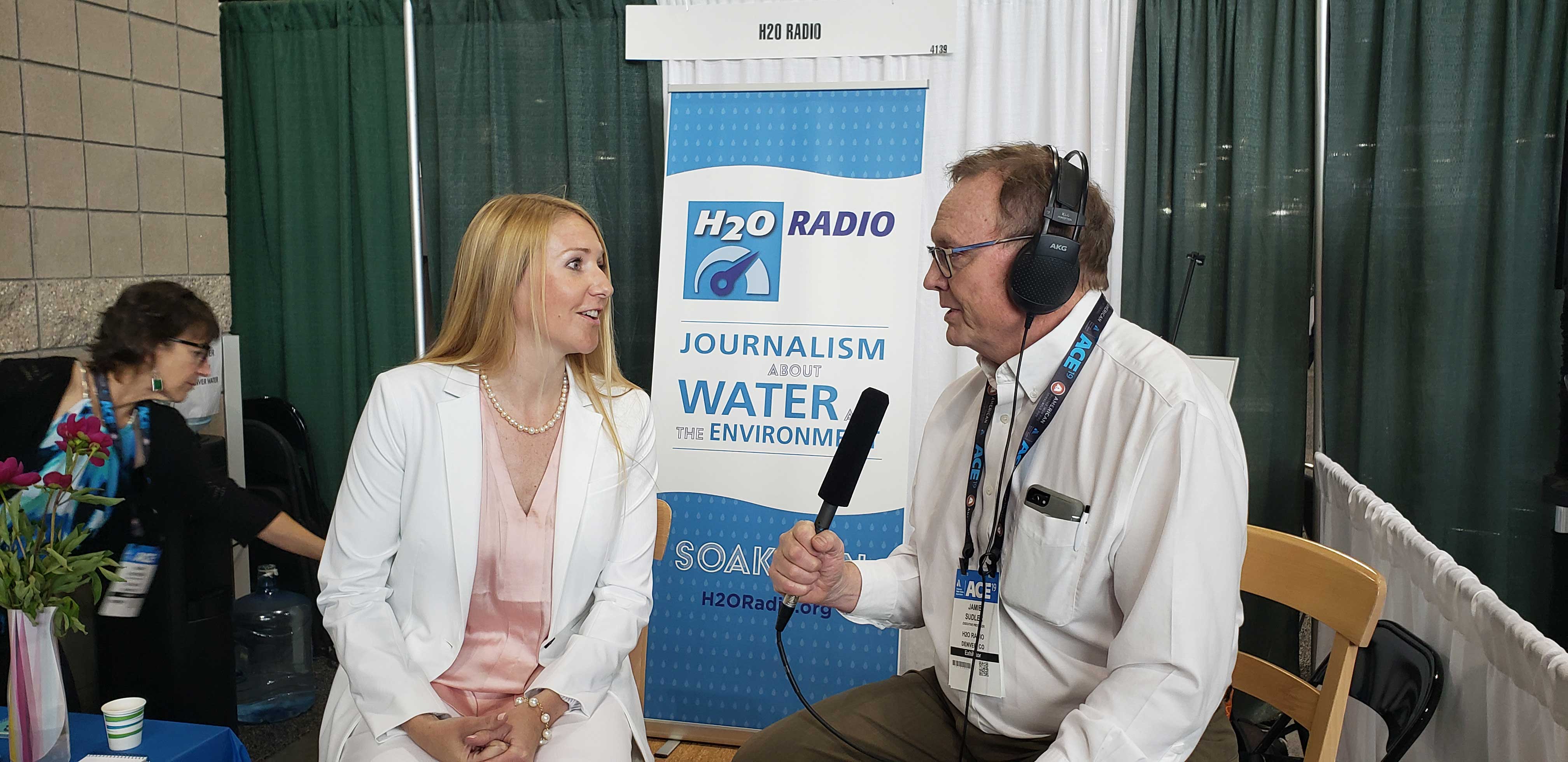 A woman talks with a man who is wearing radio headphones and carryhing a large microphone. IN the background is hte banner: H20 Radio, Journalism about Water and the Environment.