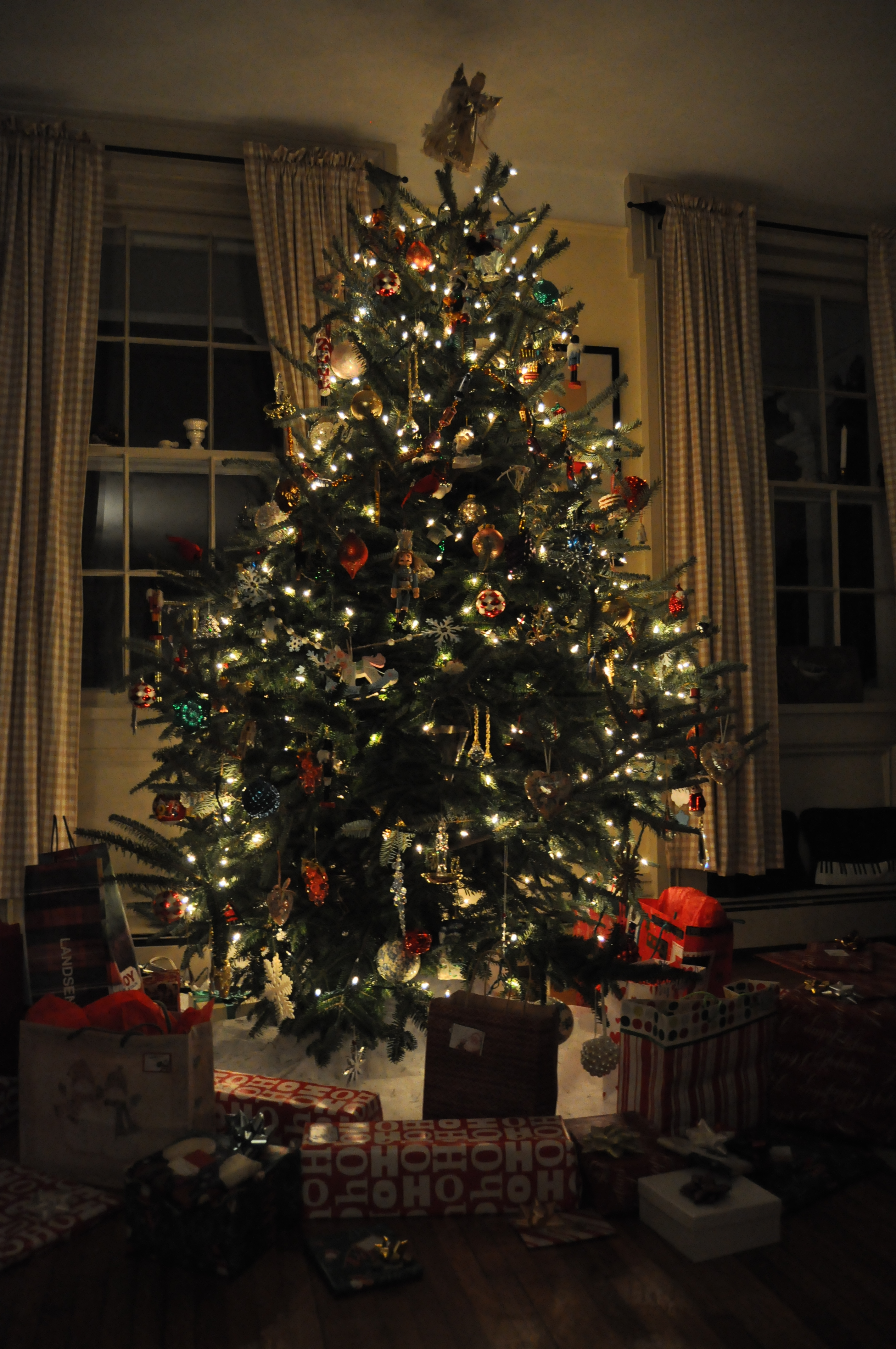 A Christmas tree in a living room is decorated with lights and ornaments with presents on the floor around it.