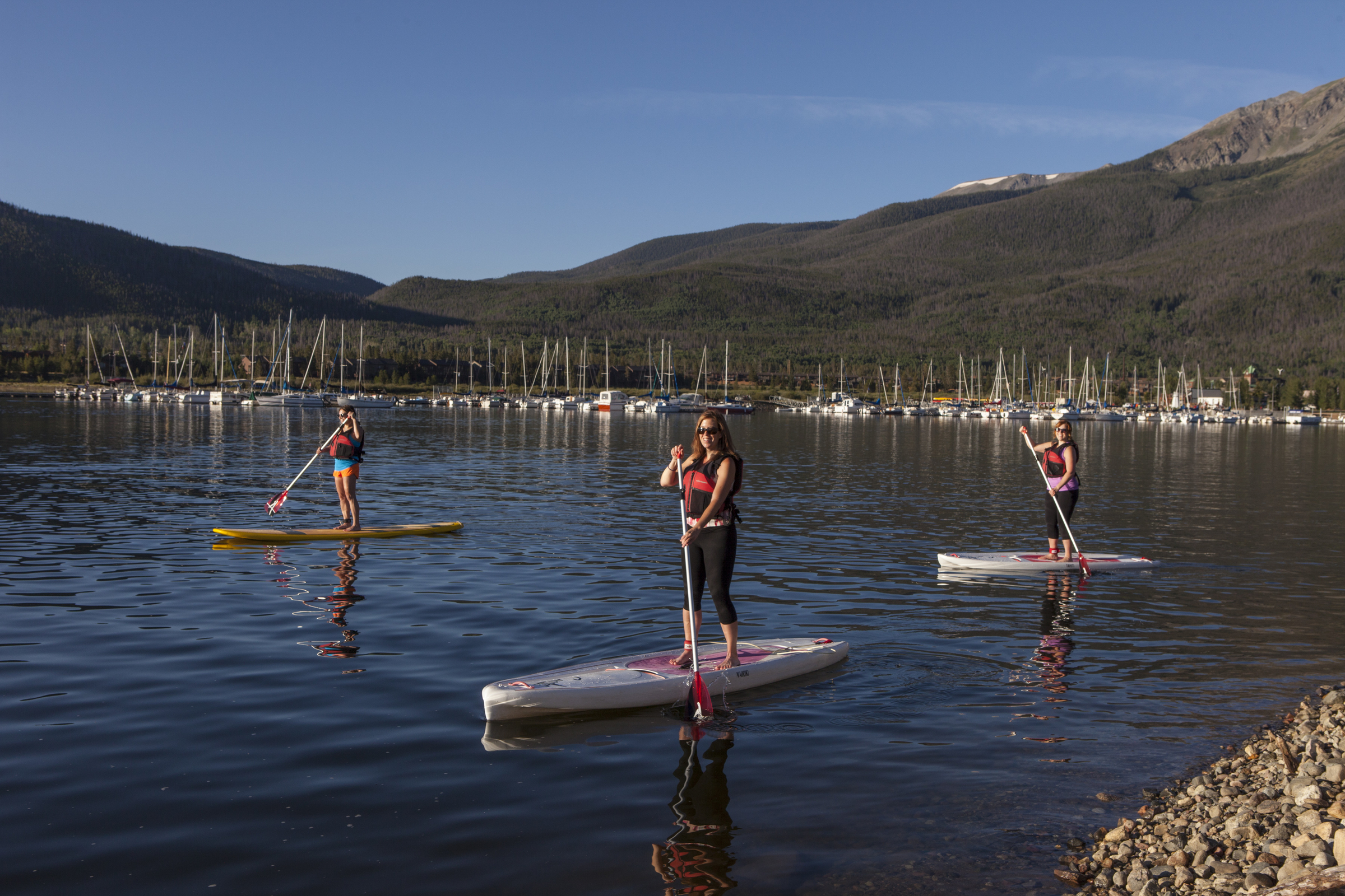 Three women on paddle boards with sail boats docked in the background.