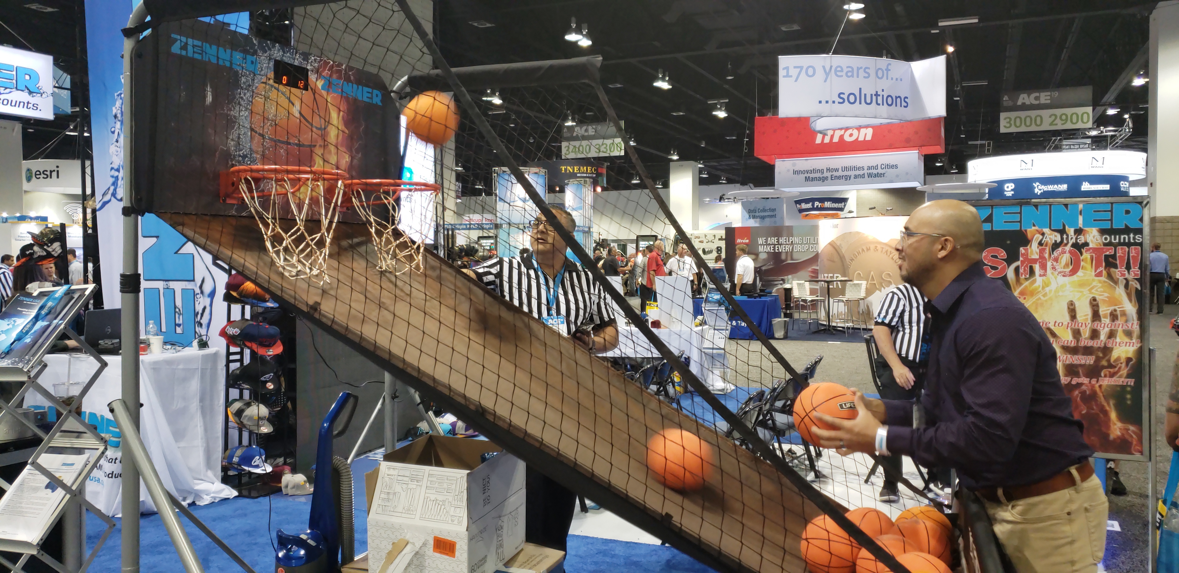 A man shoots small basketballs at two hoops on a crowded exhibit floor.