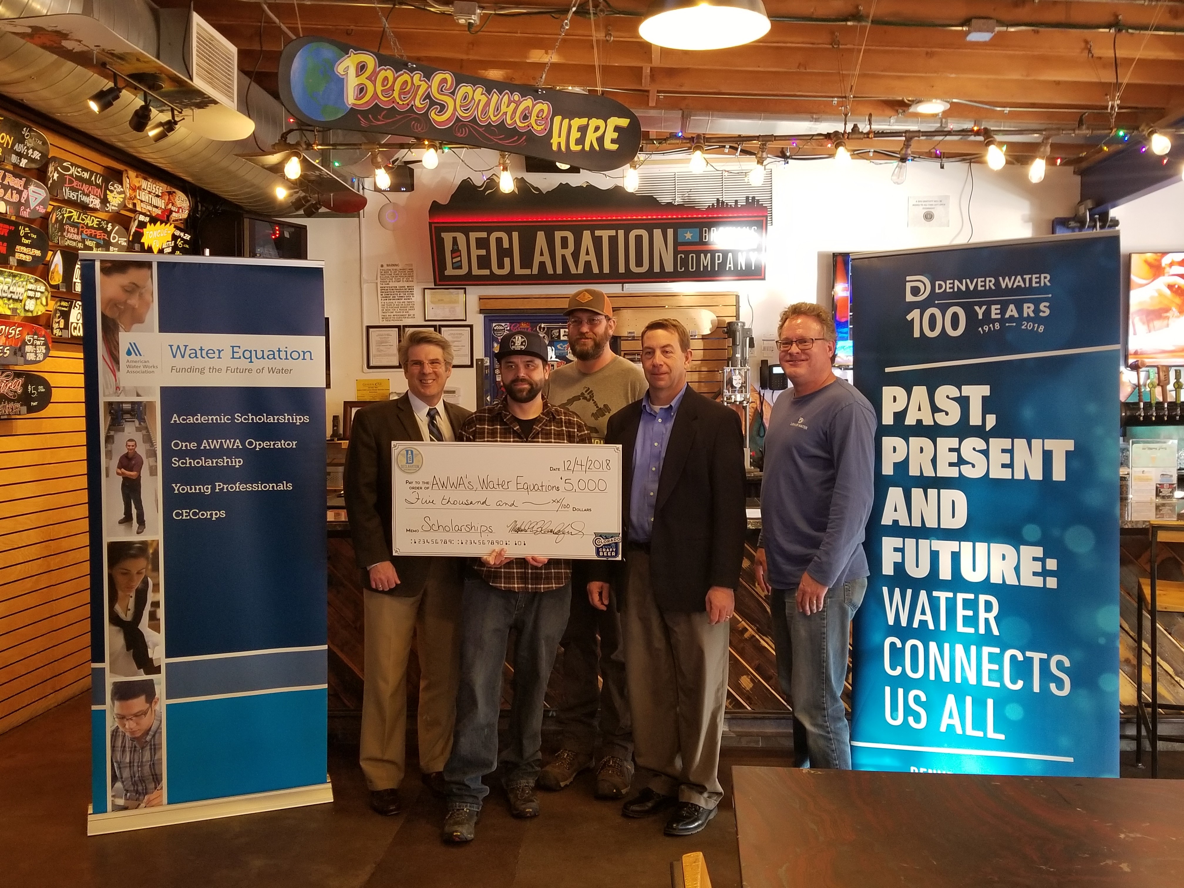 Five men standing in a bar, one of them holding a large check for $5,000, surrounded by banners from the American Water Works Association, Denver WAter, and underneath a sign for Declaration Brewery.