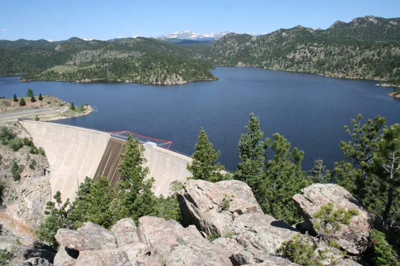 A dam is on the left of the image, with a blue body of water stretching into the distance. Trees line the shors and the sun is in the sky.