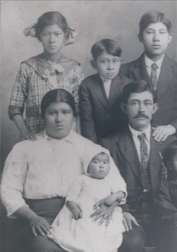 In a black and white photo, a man in a suit and tie and his wife in a white blous and hair pulled back in a bun, sit with three children, two boys in suits and ties and a girl in pigtails, are standing behind them and a baby is held on the mother's lap.