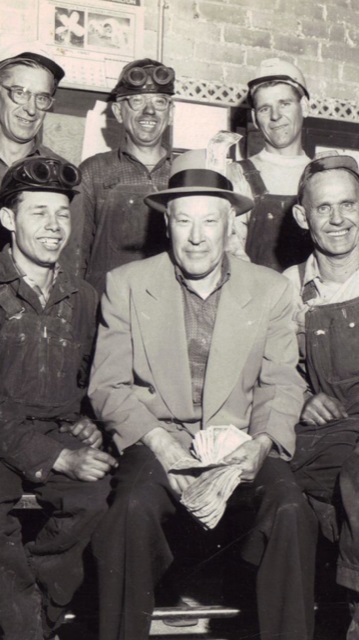 A man in a suit jacket and hat sits surrounded by other men who are wearing overall, and have goggles and hard hats on. All of them are smiling.