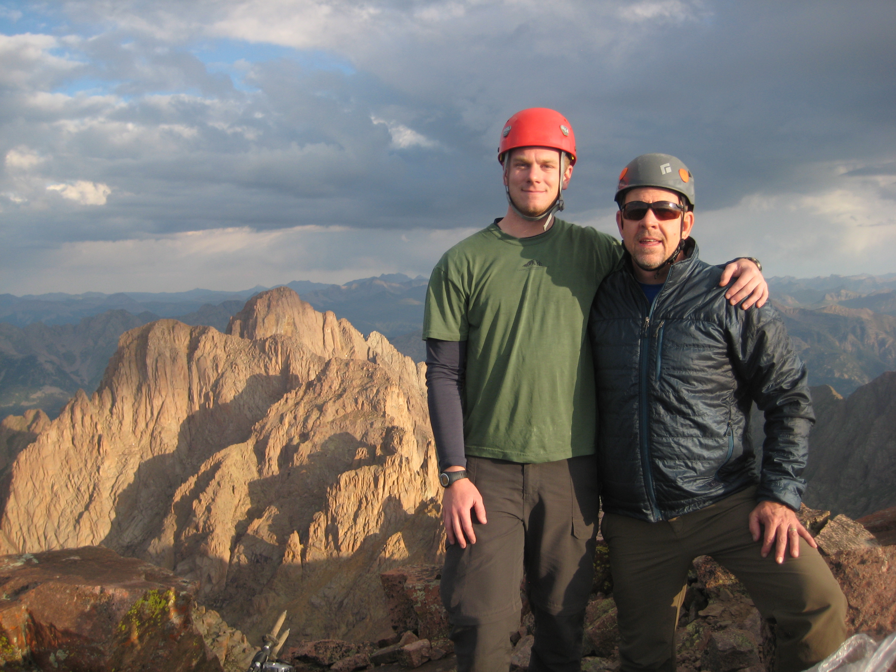 An older man and a younger man stand facing the camera, with their arms around each other, in the background looms a jagged peak under cloudy skies.