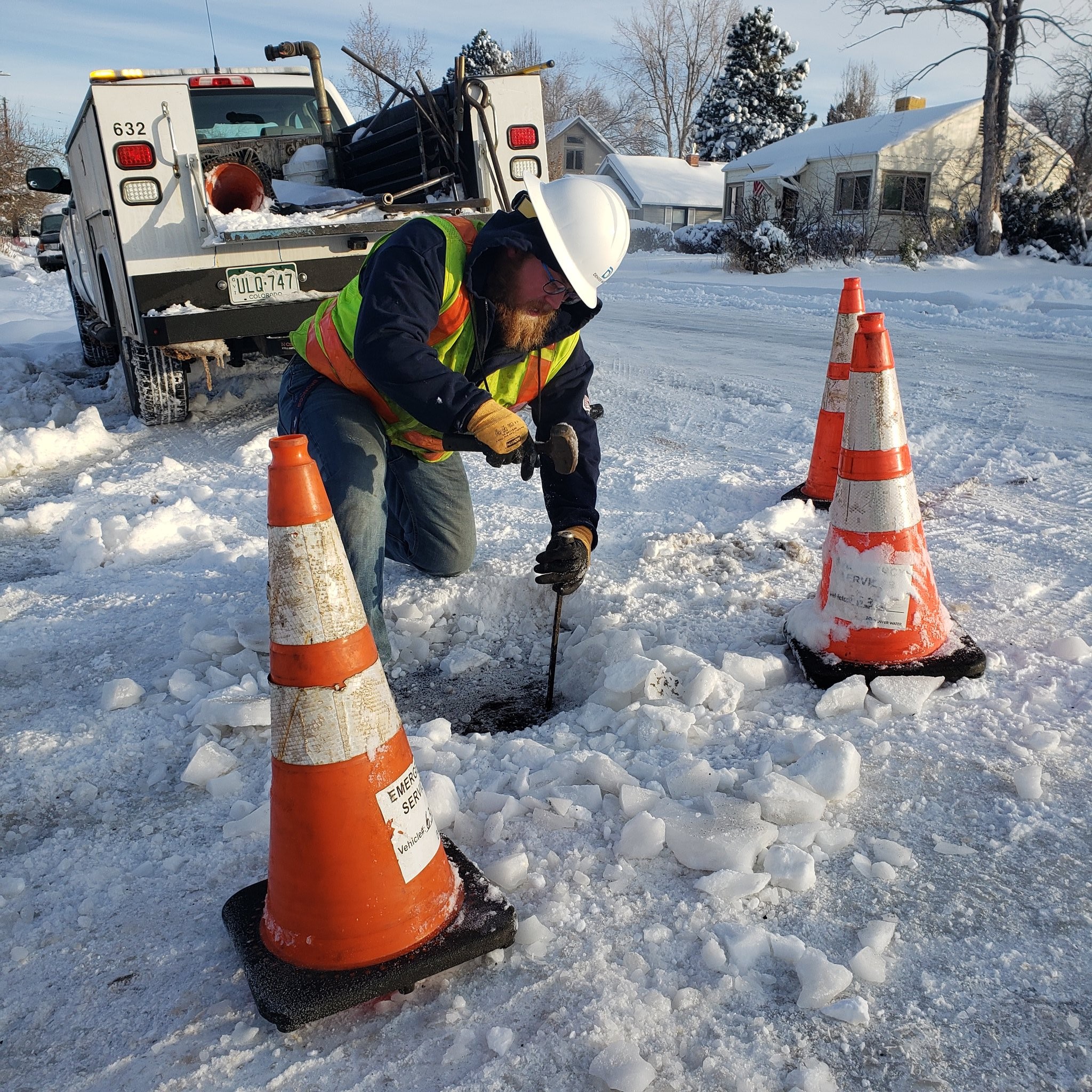 Denver Water employee is chiseling through ice to expose a shut-off valve cover. Safety cones sit around him.