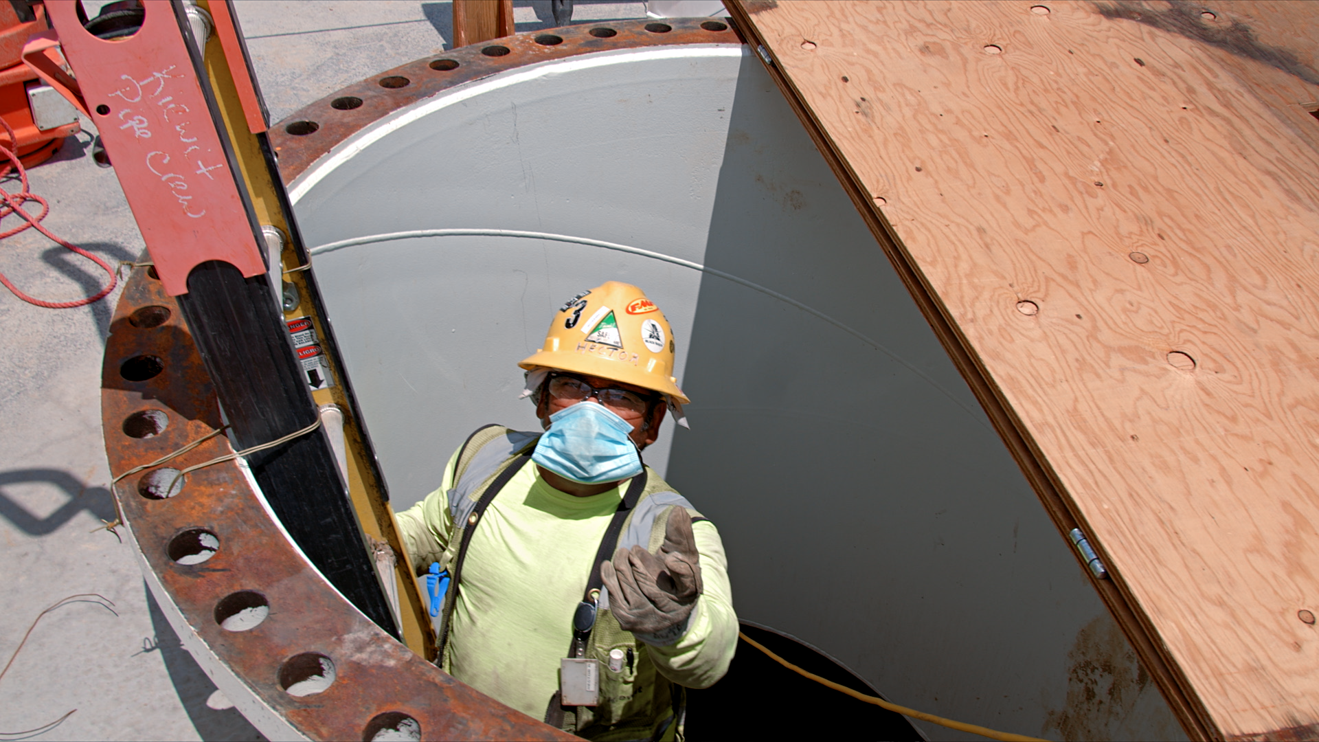 a man wearing a yellow hard hat and glasses and a blue mask climbs out of a pipe.