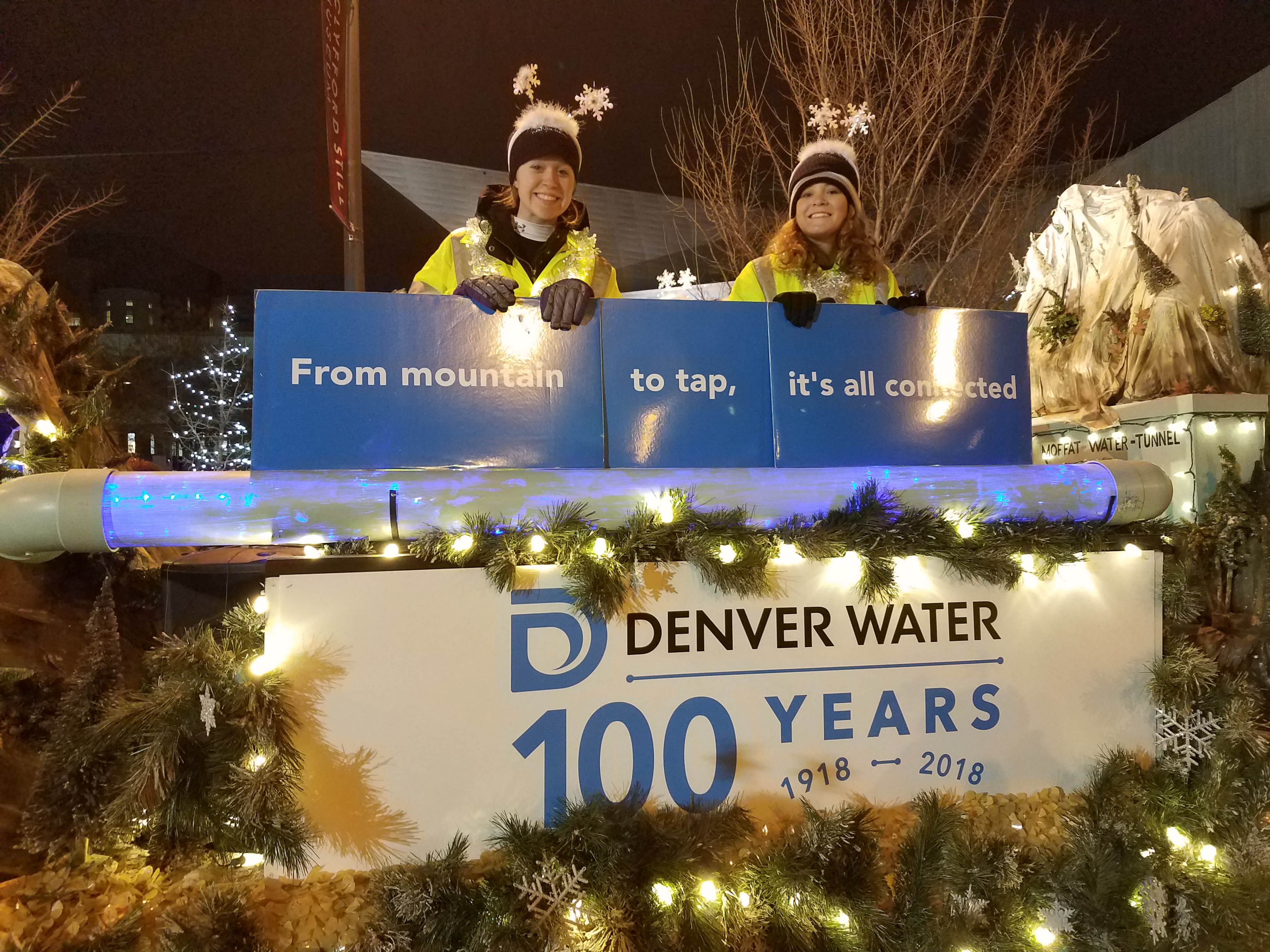 A closer look at the banner on the float saying "Denver Water, 100 years, 1918-2018). Two vwomen, smiling, hold signs that read "From mountain to tap, it's all connected."