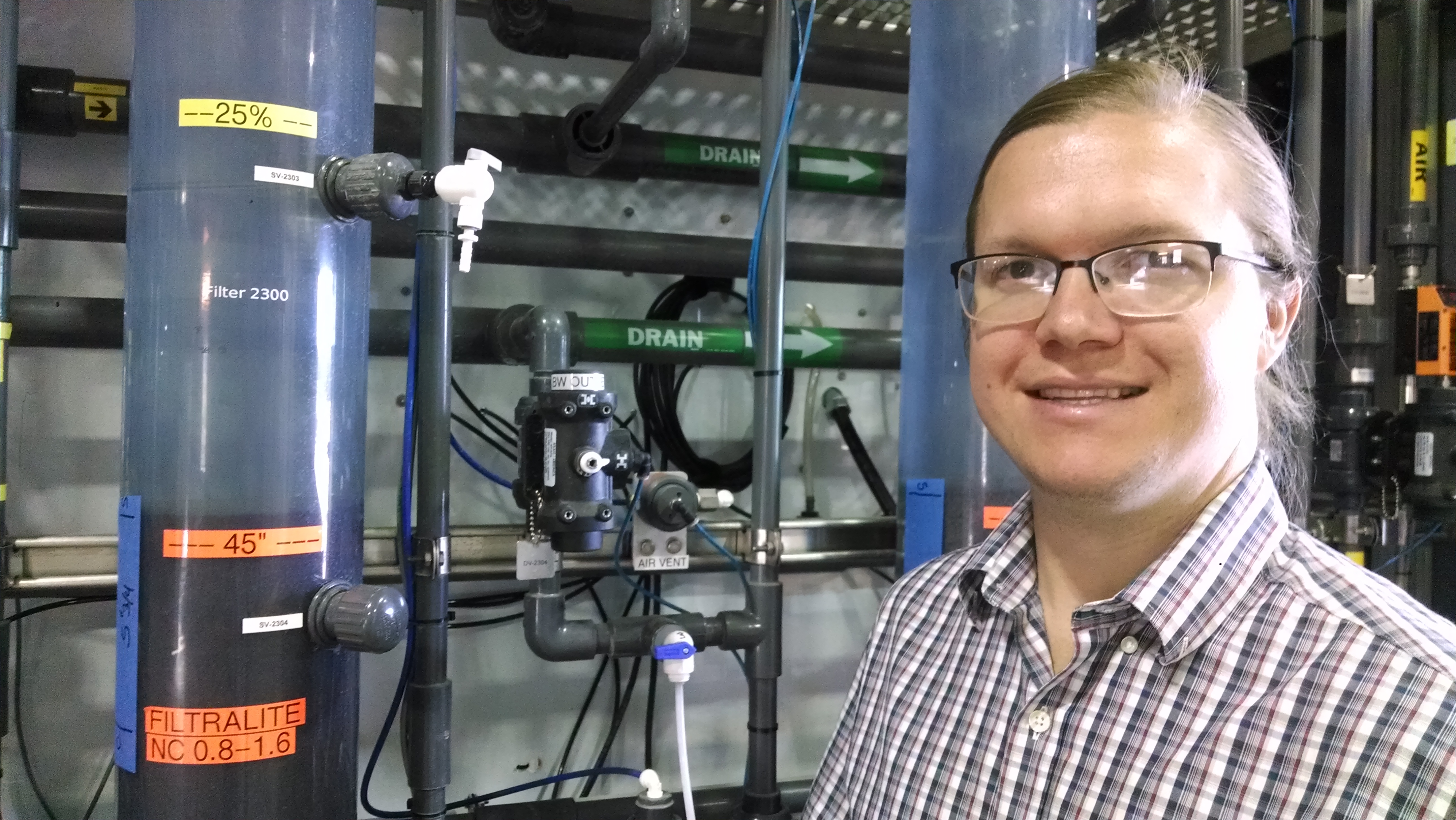 Matt Bolt is smileing for the camera next to a miniature, test treatment plant, which looks like a cylinder of water filled halfway with particles and halfway with sand.