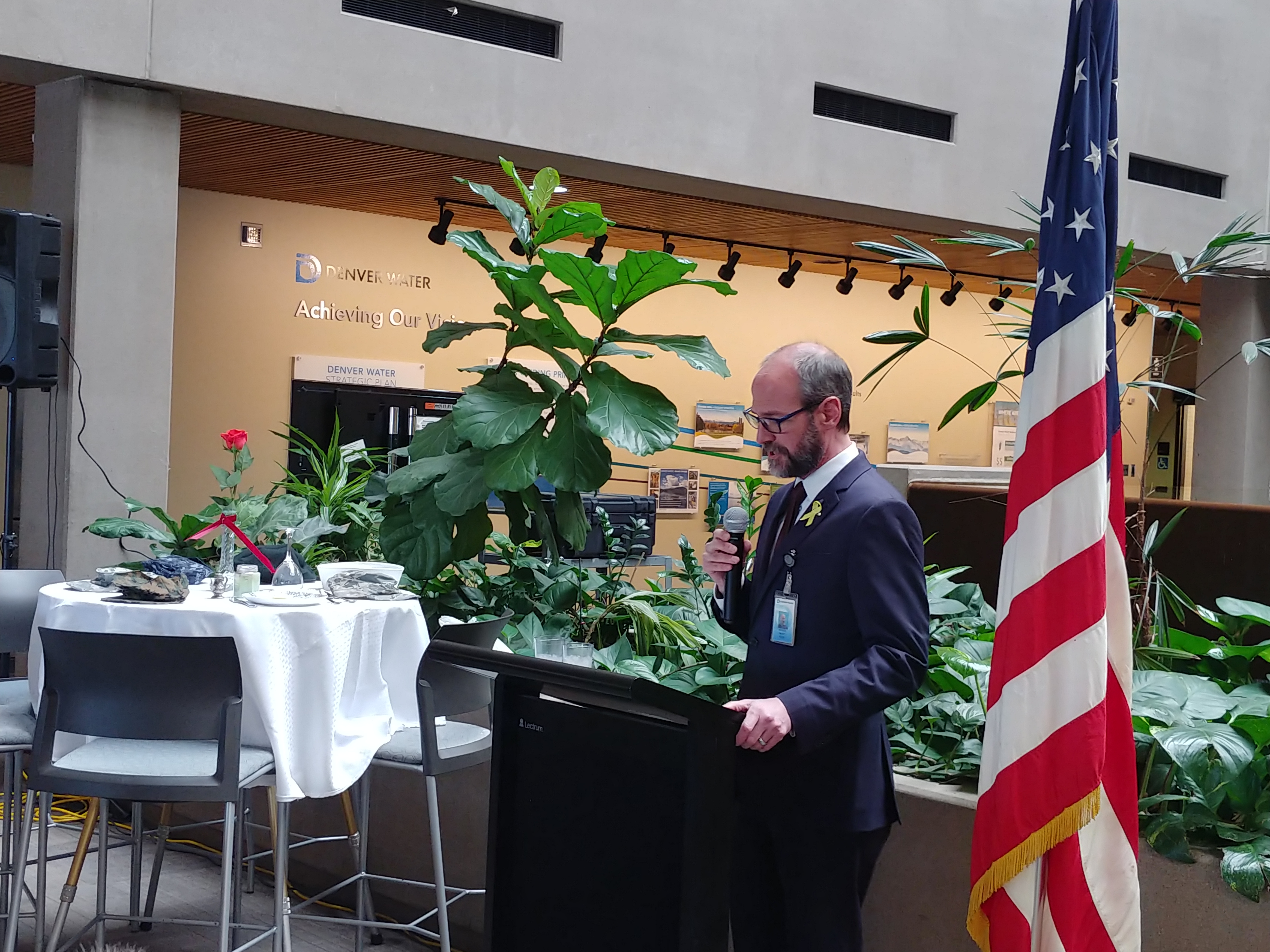 A man stands at a podium with a microphone. On his left is the U.S. flag and on his right is the "missing man" table.
