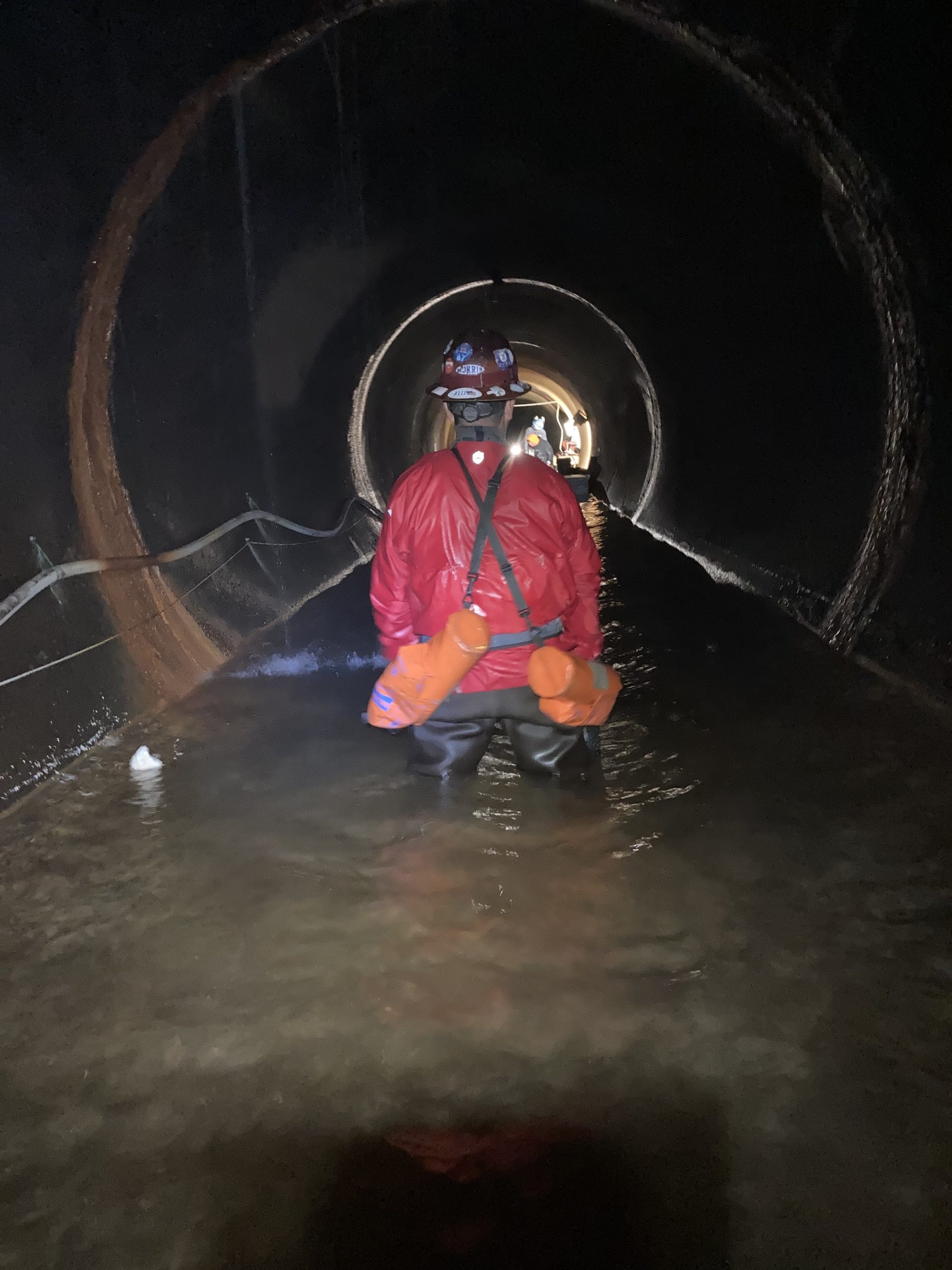 a man stands in a tunnel, his back to the camera, wearing a hard hat, and other gear and water up above his knees.