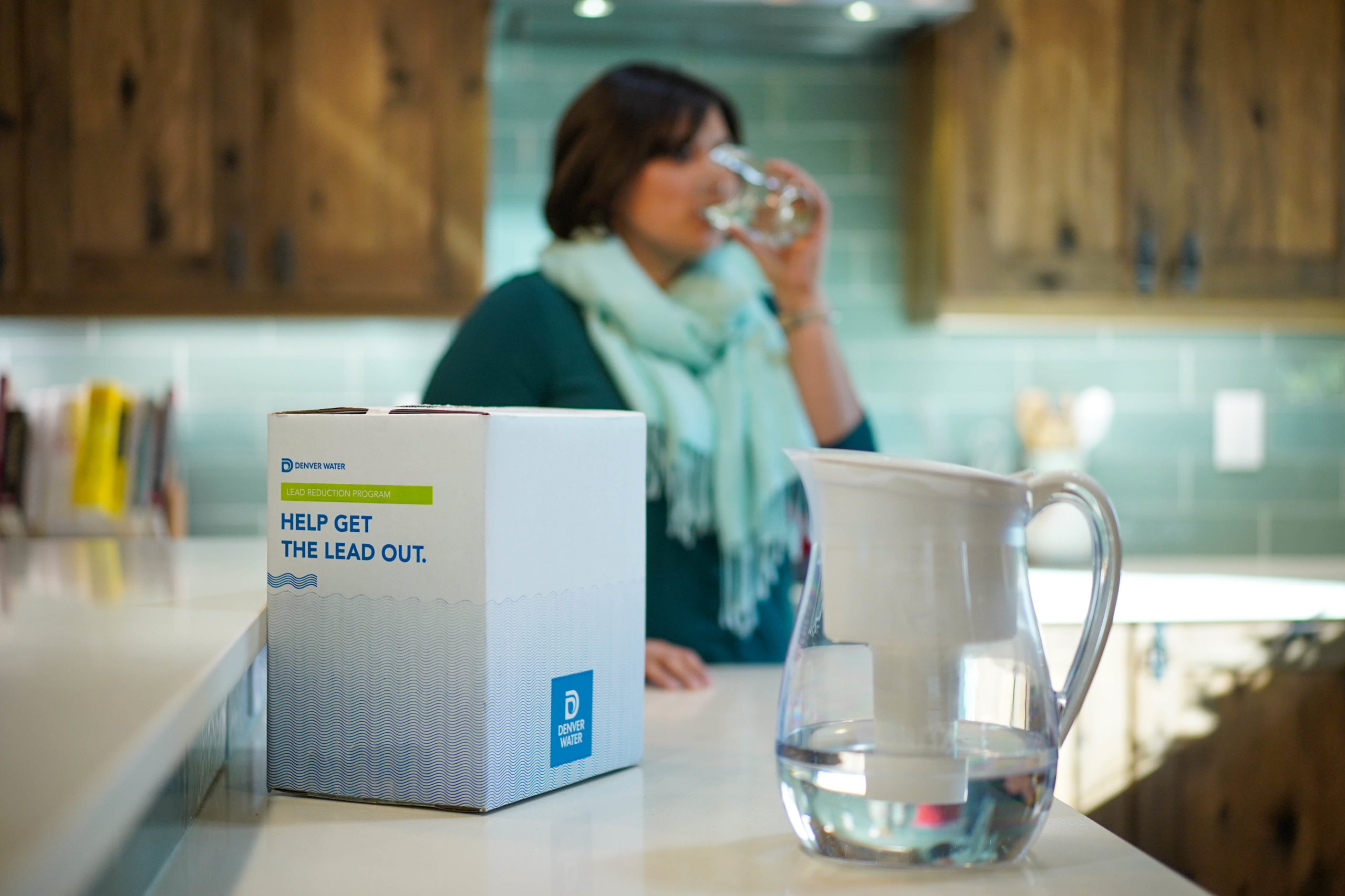 A woman drinks water from a glass, in the foreground is a box and water pitcher with a filter.