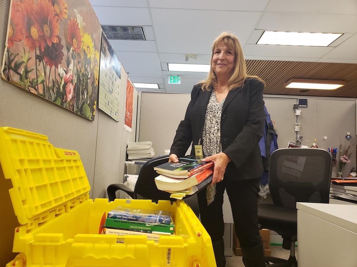 A woman holds a stack of books to put them into a yellow bin.