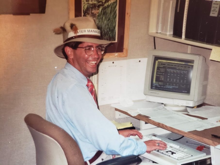 A man smiles for the campera with a fedora and a huge computer on his desk.