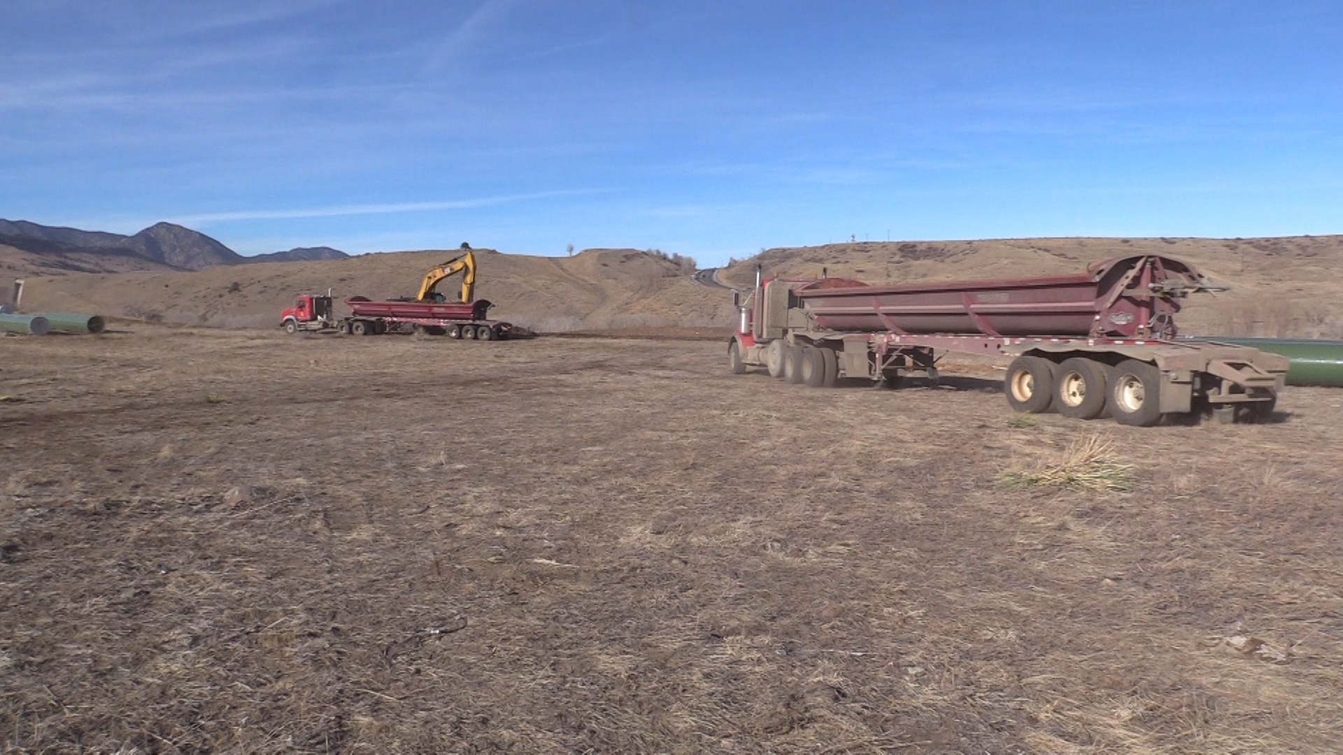 Red trucks are lined up to be filled with dirt dug from the earth. In the distance, Highway 93 carves through the foothills under a blue sky.