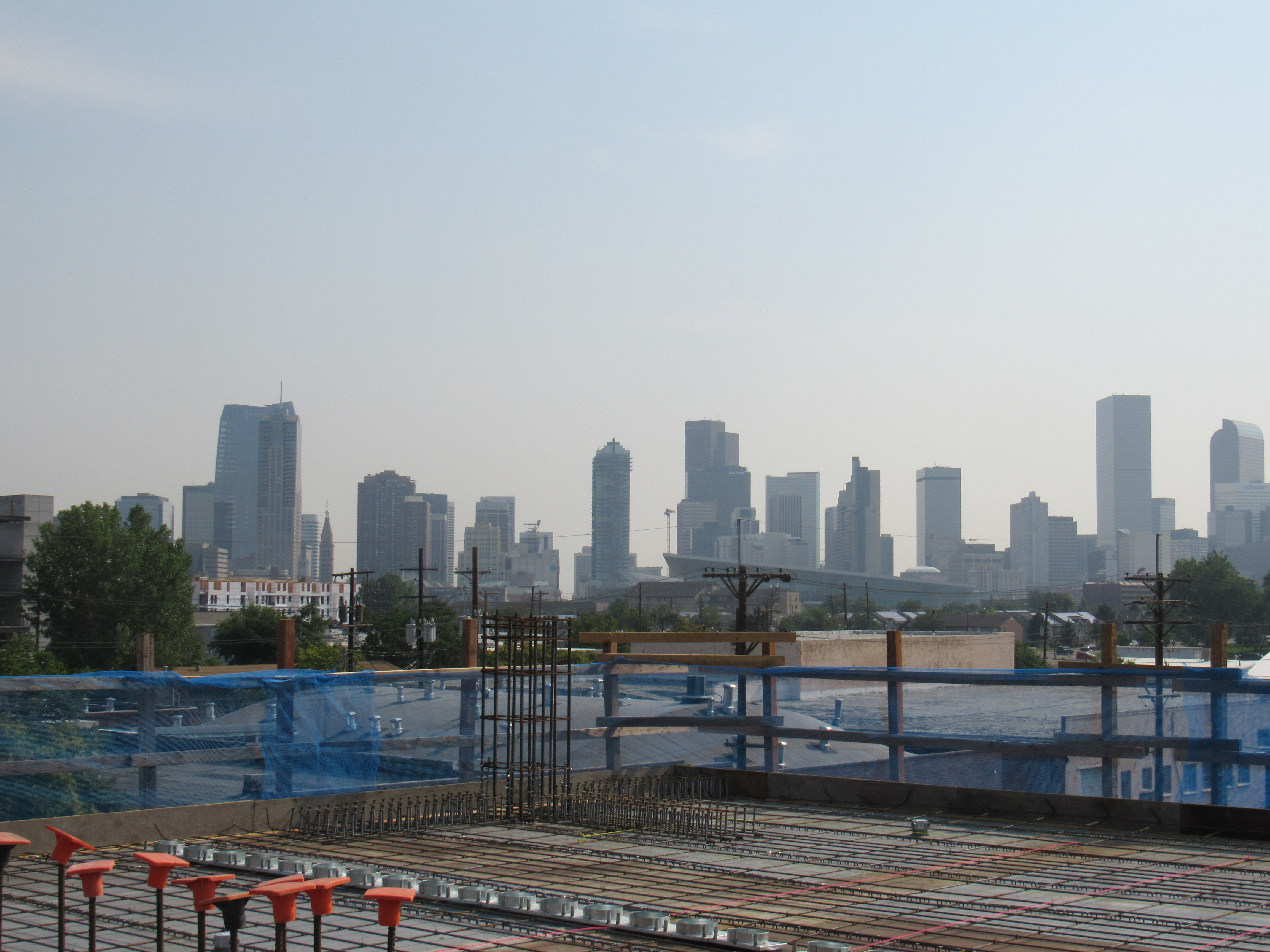 Foregrond shows the roof, under construction, with the downtown Denver skyline in the dstance under a hazy sky.