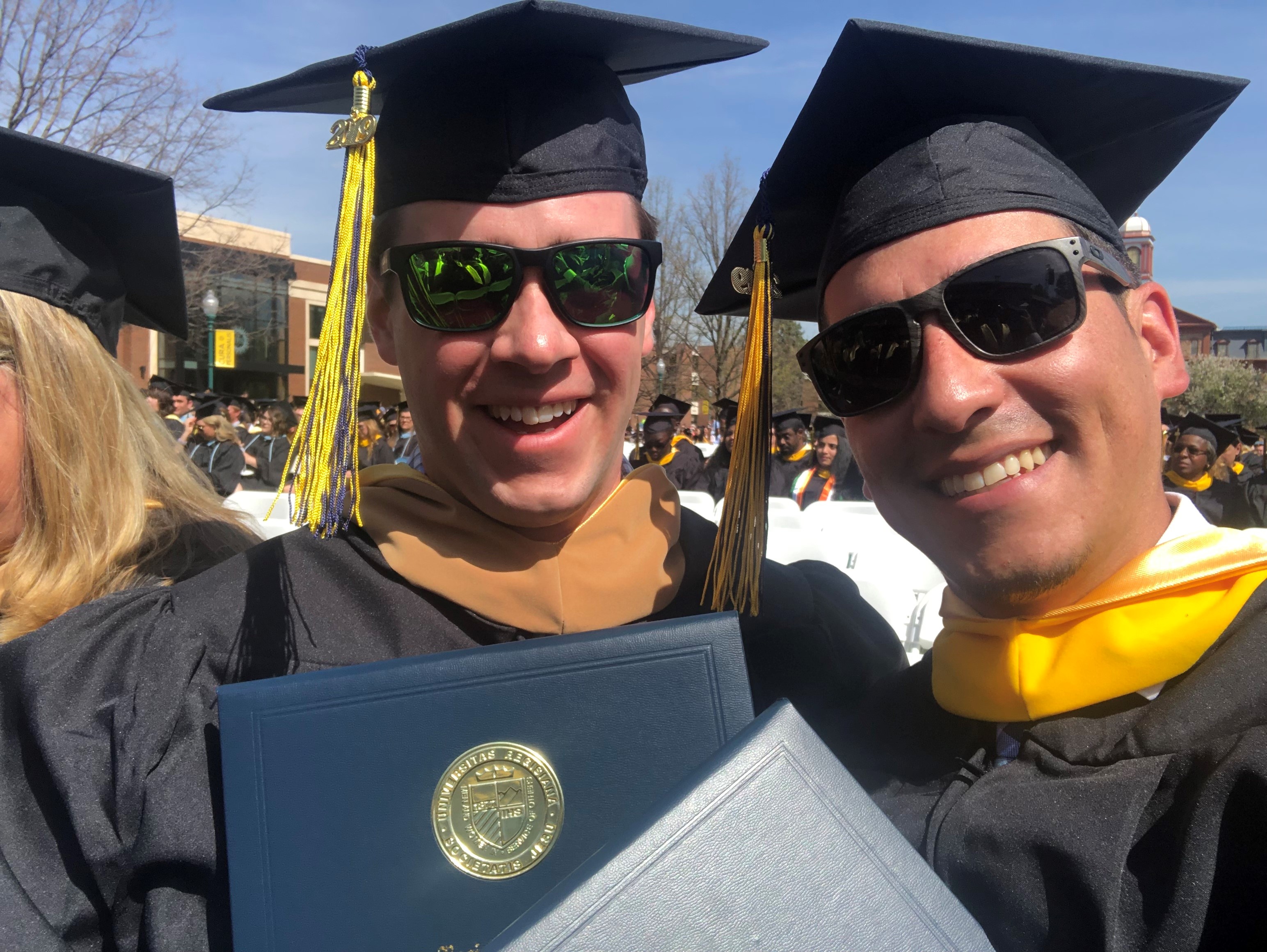 Two men in sunglasses with graduation caps, tassels and gowns smile for the camera.